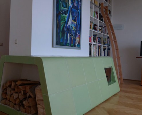 Kachelofen Wien 2: Liegender grüner Ofenkörper mit integriertem Holzlager und Sichtfenster, Kacheln mittels eigens angefertigter Holzform gebaut, abgerundete und etwas auskragende Form.